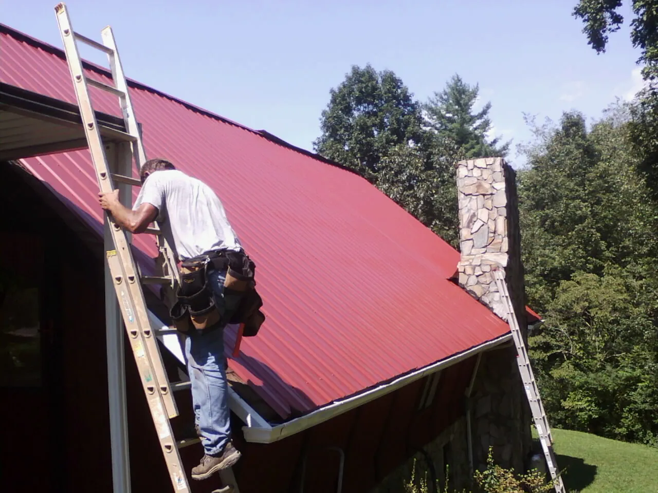 Roofer on a ladder beside a red metal roof and stone chimney — Soffit & Fascia Repair by Roofing Brothers in Penn